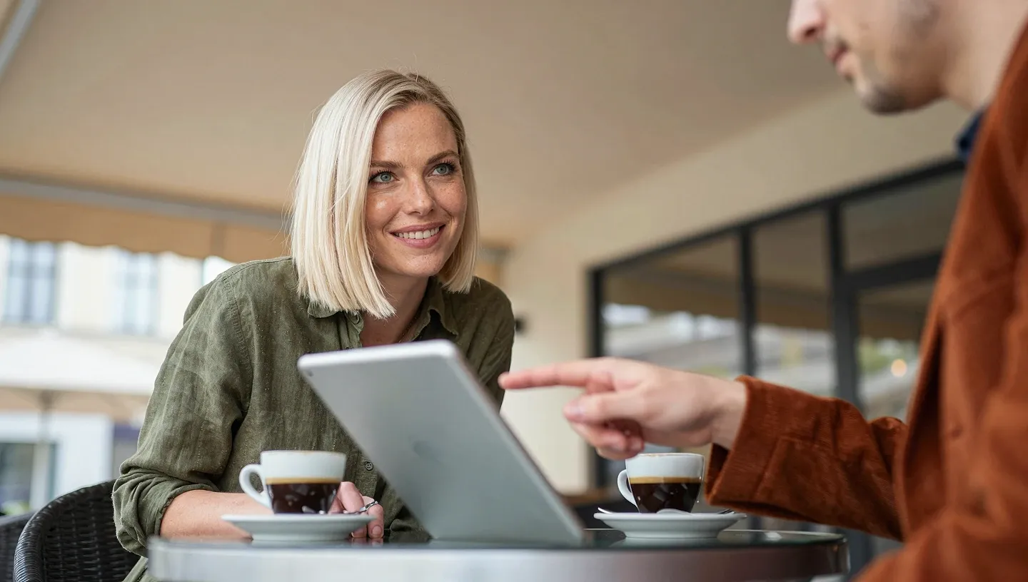 Two women at a café terrace lean over a tablet and espresso cups as they discuss SEO pricing tiers in diffused daylight