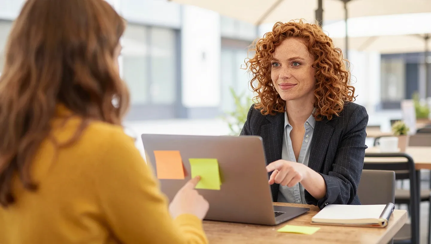 A creator with curly auburn hair and her manager meet at a café terrace with coffee, a softly glowing laptop, notebook, and colorful sticky notes