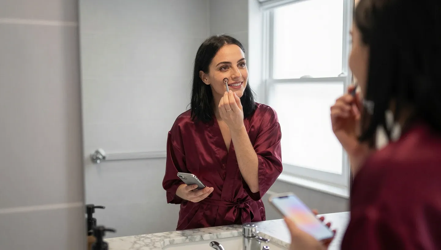 A late twenties woman in a burgundy silk robe applies makeup at a bathroom mirror while her phone is propped on the sink