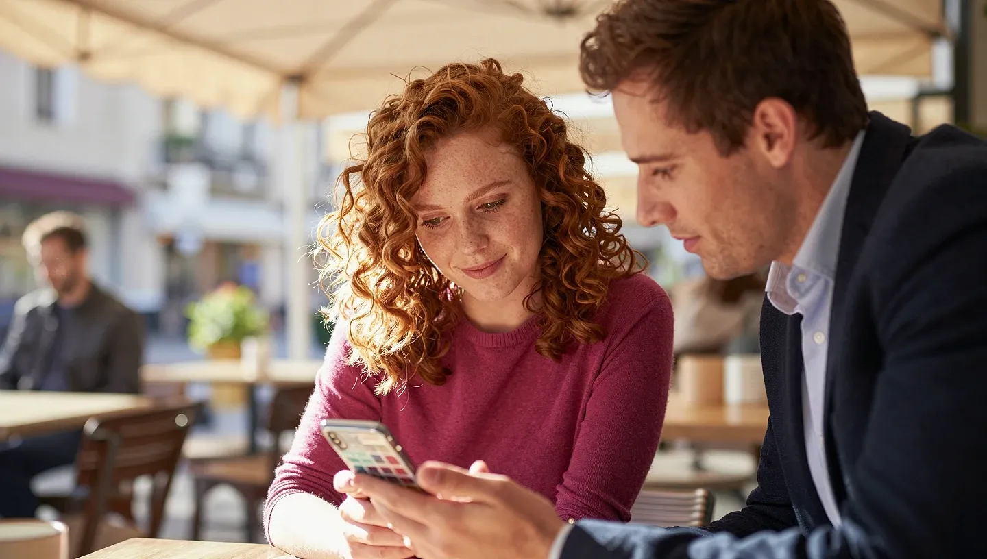 Curly auburn-haired creator and her manager glance at a smartphone with blurred calendar blocks on a sunlit café terrace