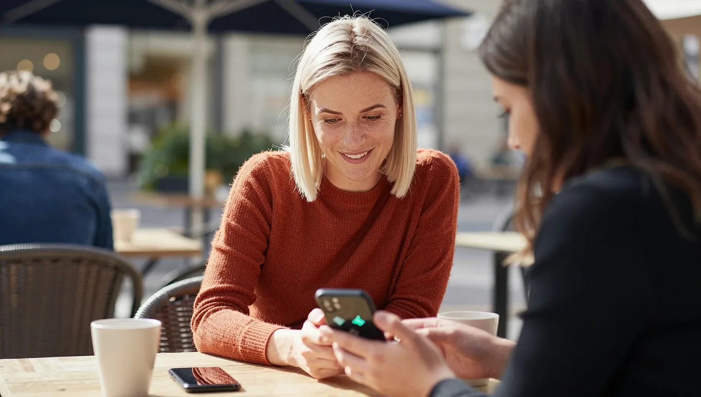 A content creator with a platinum-blonde bob and her manager review a smartphone over coffee on a sunny café terrace